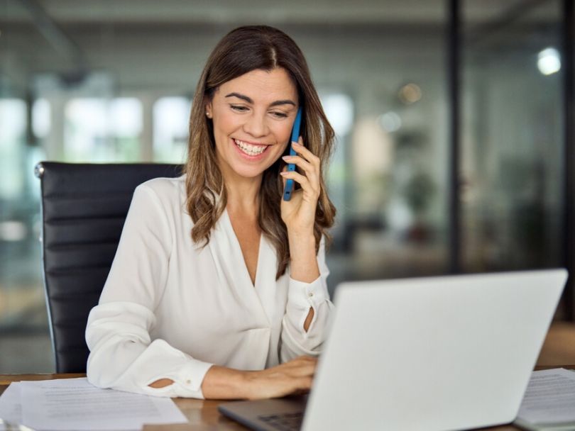 A professional woman chats on the phone while working on her laptop in an office